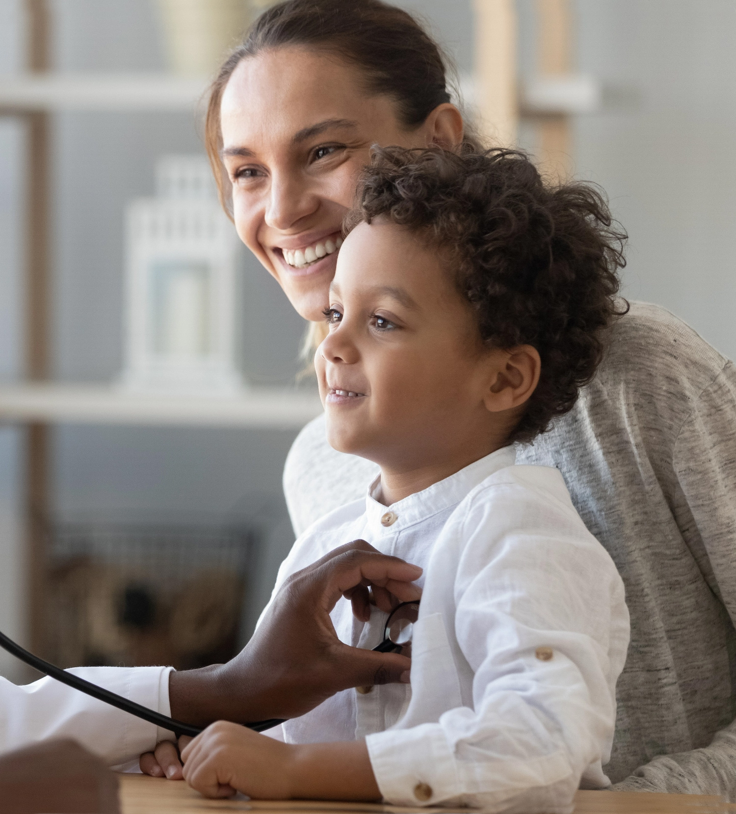 A mother with her son on her lap both smiling with doctor off camera holding the stethoscope over the young boy's heart