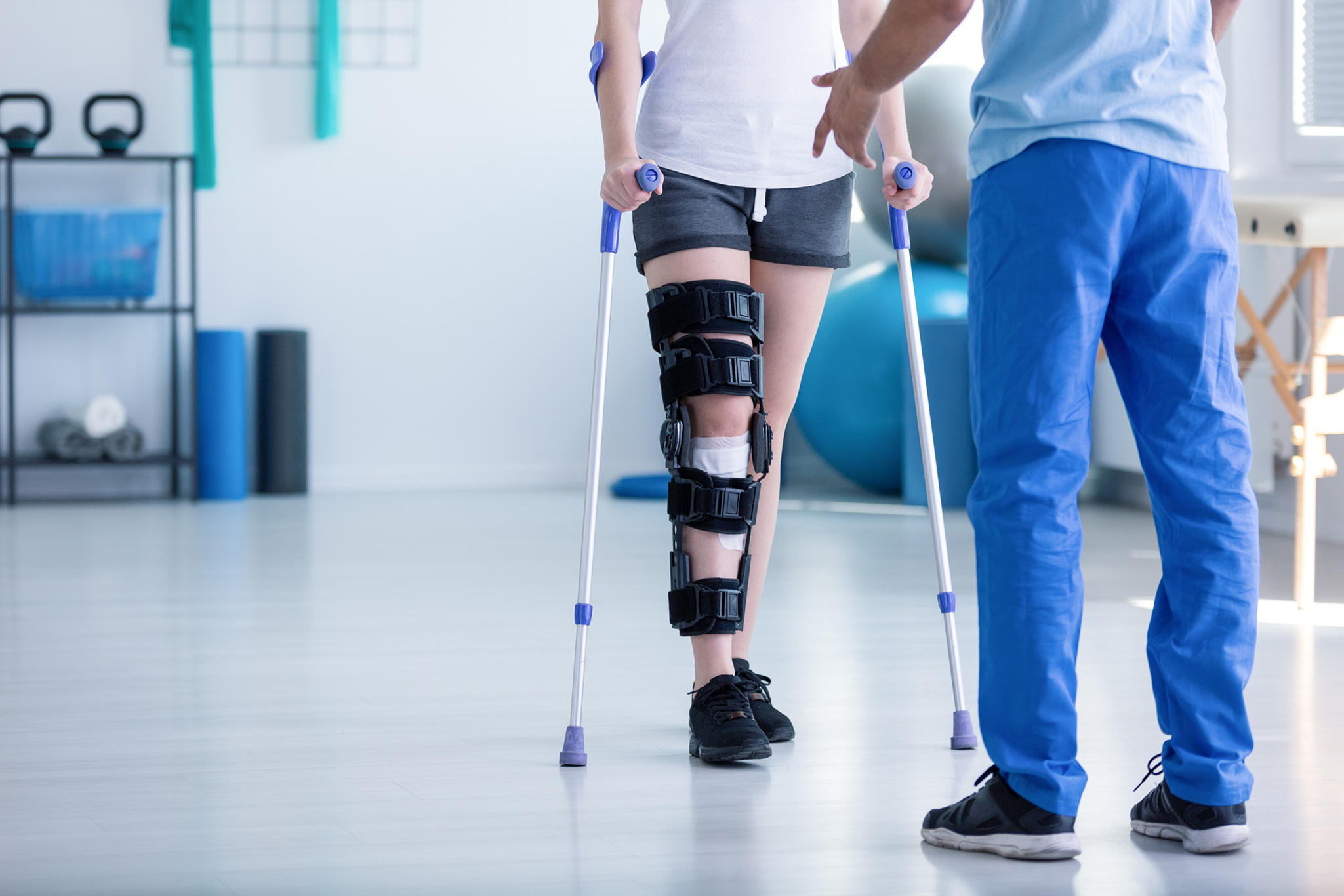 A wider angle shot of person in crutches wearing a high knee brace and bandaged knee in a sterile setting with the PT's backside in the foreground (blurred)