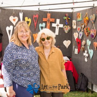 Art in the Park 2021 smiling women in booth with cross and heart art